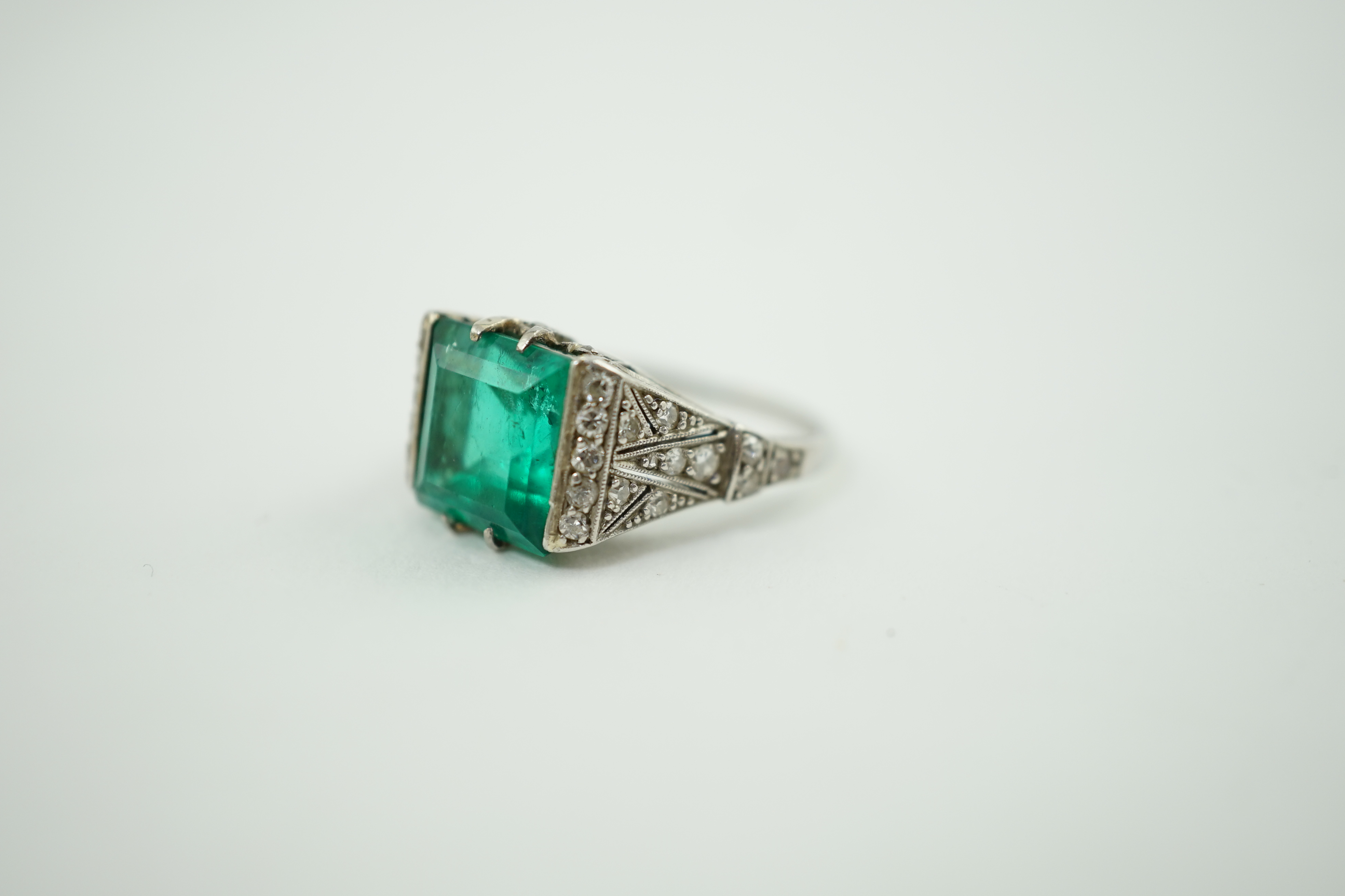 A 1920's/1930's platinum and square cut emerald set dress ring, with pierced and millegrain diamond cluster set shoulders